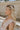 Side view of female model wearing the Emilia Satin Plunge Neckline Sleeveless Bodysuit which features Satin Fabric, Surplice Neckline with Button Snap, Sleeveless and Thong Bottom with Button Closure. the bodysuit is available in taupe and white