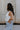 Side view of female model wearing the Emilia Satin Plunge Neckline Sleeveless Bodysuit which features Satin Fabric, Surplice Neckline with Button Snap, Sleeveless and Thong Bottom with Button Closure. the bodysuit is available in taupe and white