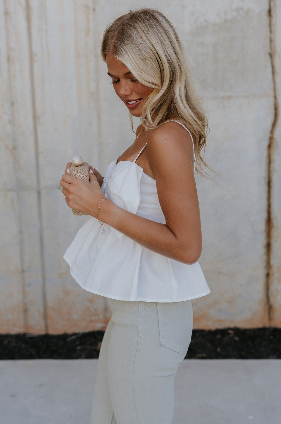 Side view of female model wearing the Alina Off White Bow Poplin Tank which features White Lightweight Fabric, Cropped Waist, Front Oversized Bow Detail, Adjustable Straps and Monochrome Back Zipper with Hook Closure