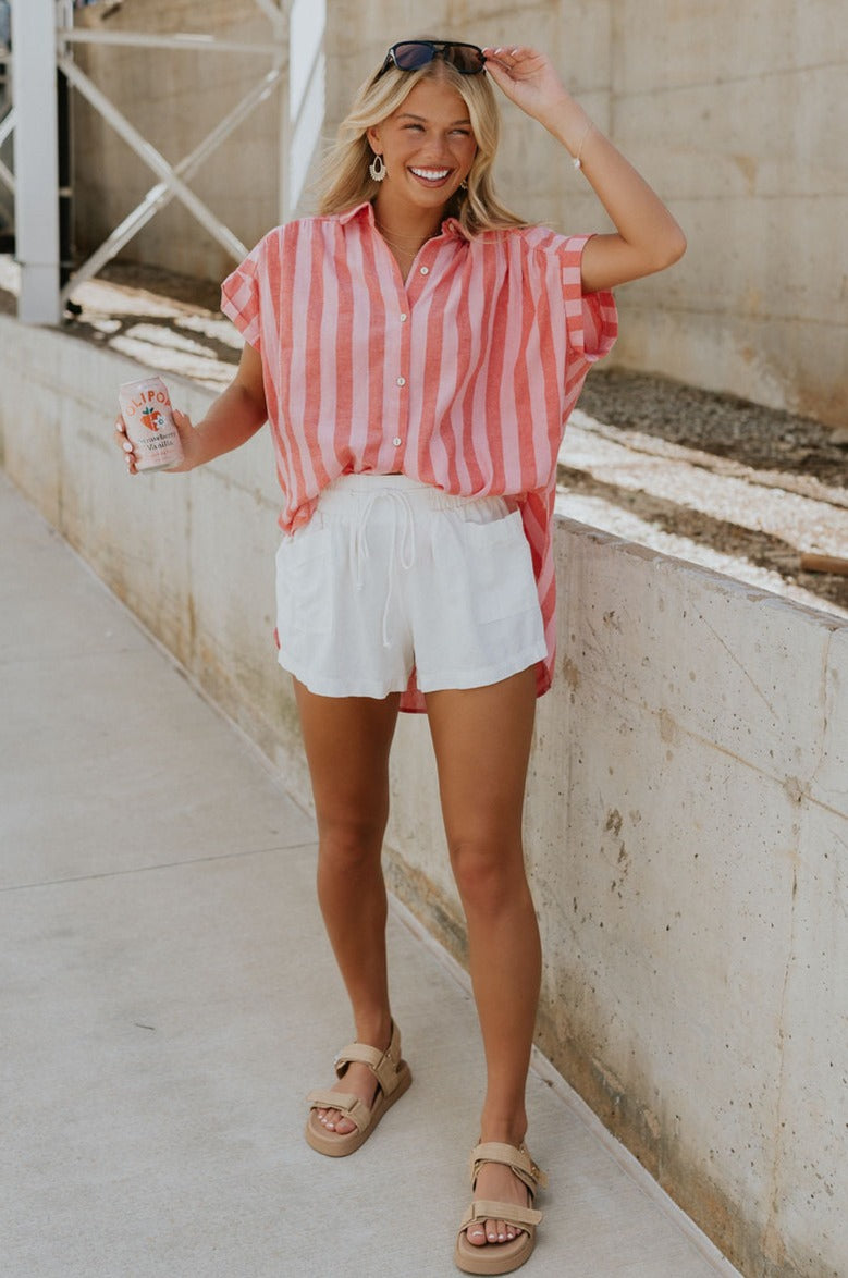 Full body view of female model wearing the Sienna Pink & Red Stripe Button-Up Short Sleeve Top which features Red and Pink Stripe Pattern, Scooped Hem, Slit Details on each side, Wooden Front Button-Up, Collared Neckline and Short Sleeves