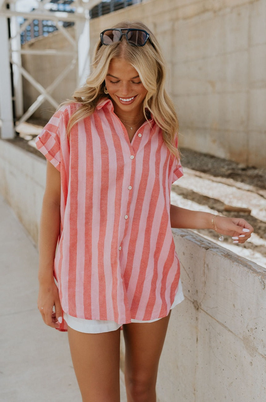 front view of female model wearing the Sienna Pink & Red Stripe Button-Up Short Sleeve Top which features Red and Pink Stripe Pattern, Scooped Hem, Slit Details on each side, Wooden Front Button-Up, Collared Neckline and Short Sleeves