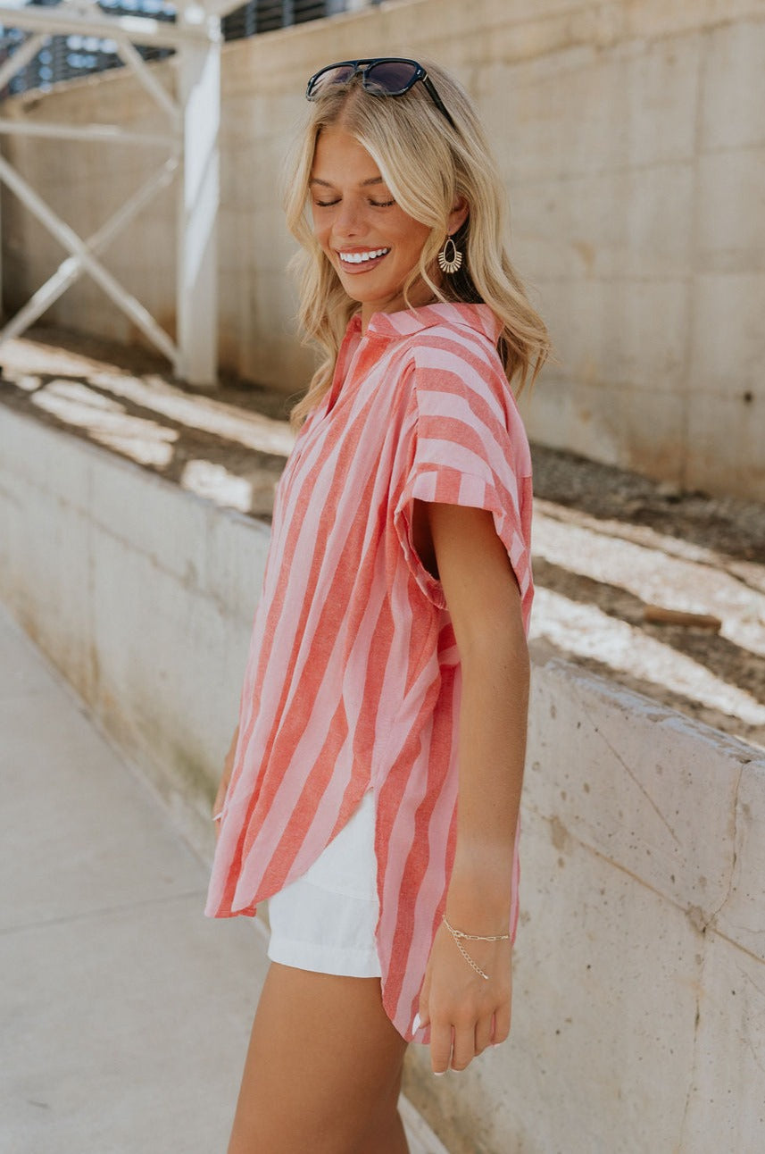 Side view of female model wearing the Sienna Pink & Red Stripe Button-Up Short Sleeve Top which features Red and Pink Stripe Pattern, Scooped Hem, Slit Details on each side, Wooden Front Button-Up, Collared Neckline and Short Sleeves