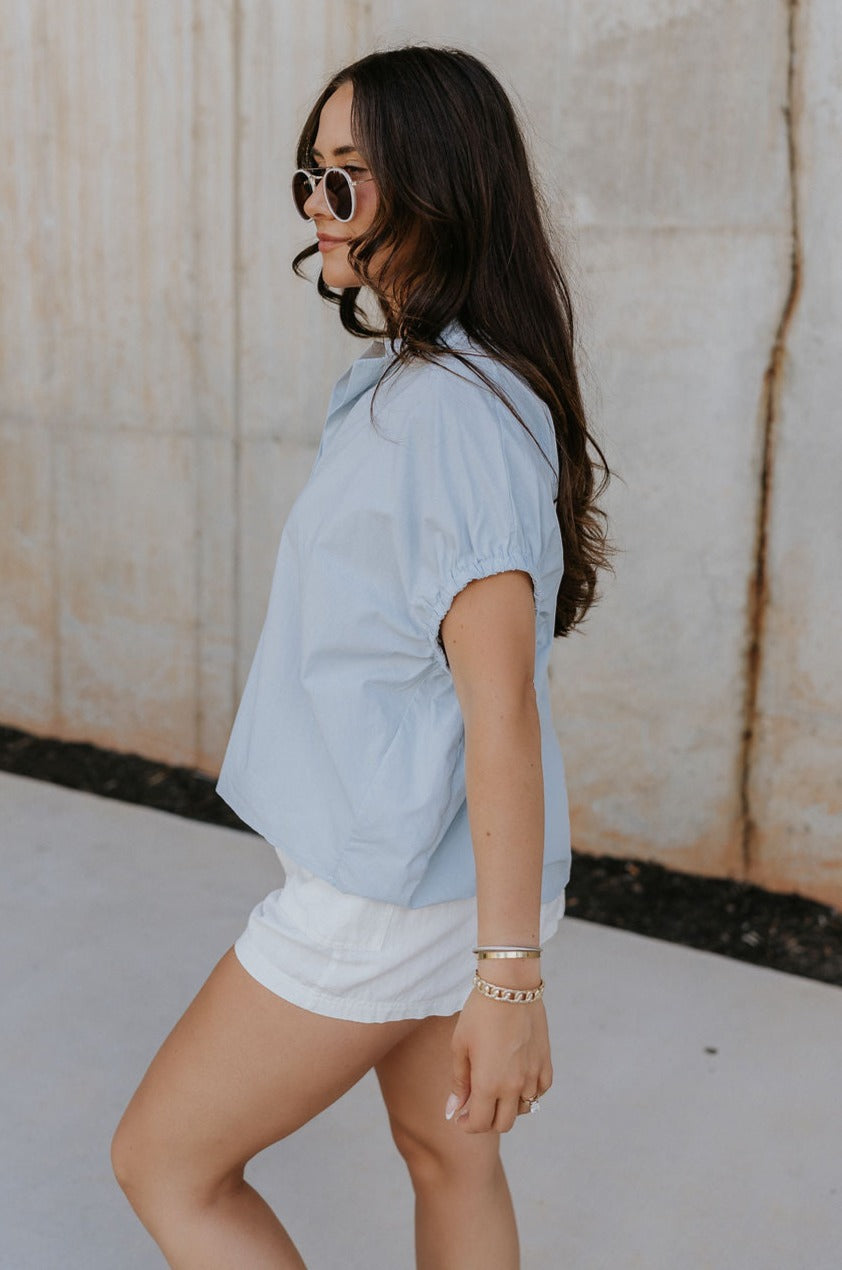 Side view of female model wearing the Noelle Chambray Blue Short Sleeve Top which features Light Blue Cotton Fabric, Slight Cropped Waist, Short Puff Sleeves and V-Neckline
