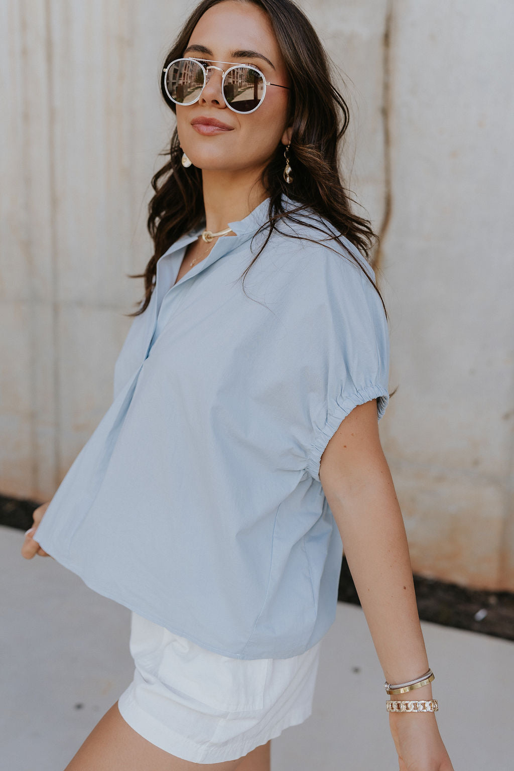 Side view of female model wearing the Noelle Chambray Blue Short Sleeve Top which features Light Blue Cotton Fabric, Slight Cropped Waist, Short Puff Sleeves and V-Neckline