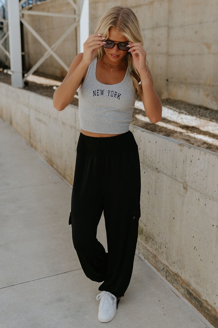 Full body view of female model wearing the Serena Black Cargo Drawstring Pants which features Black Lightweight Fabric, Jogger Pant Legs with Drawstring Ties, Cargo Pockets with Tortoise Buttons and Smocked Elastic Waistband
