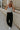 Full body view of female model wearing the Serena Black Cargo Drawstring Pants which features Black Lightweight Fabric, Jogger Pant Legs with Drawstring Ties, Cargo Pockets with Tortoise Buttons and Smocked Elastic Waistband