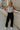 Full body view of female model wearing the Serena Black Cargo Drawstring Pants which features Black Lightweight Fabric, Jogger Pant Legs with Drawstring Ties, Cargo Pockets with Tortoise Buttons and Smocked Elastic Waistband
