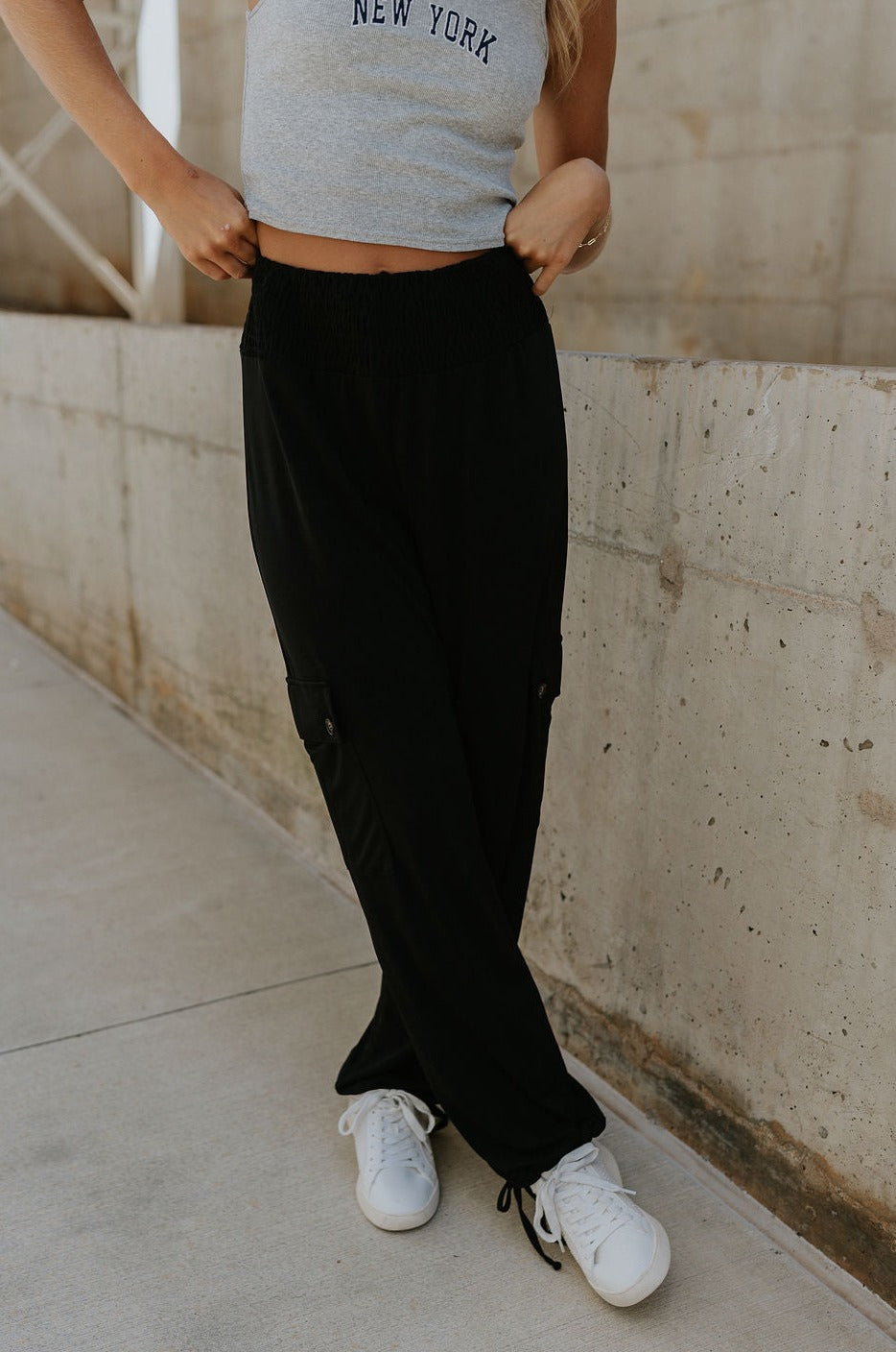 front view of female model wearing the Serena Black Cargo Drawstring Pants which features Black Lightweight Fabric, Jogger Pant Legs with Drawstring Ties, Cargo Pockets with Tortoise Buttons and Smocked Elastic Waistband