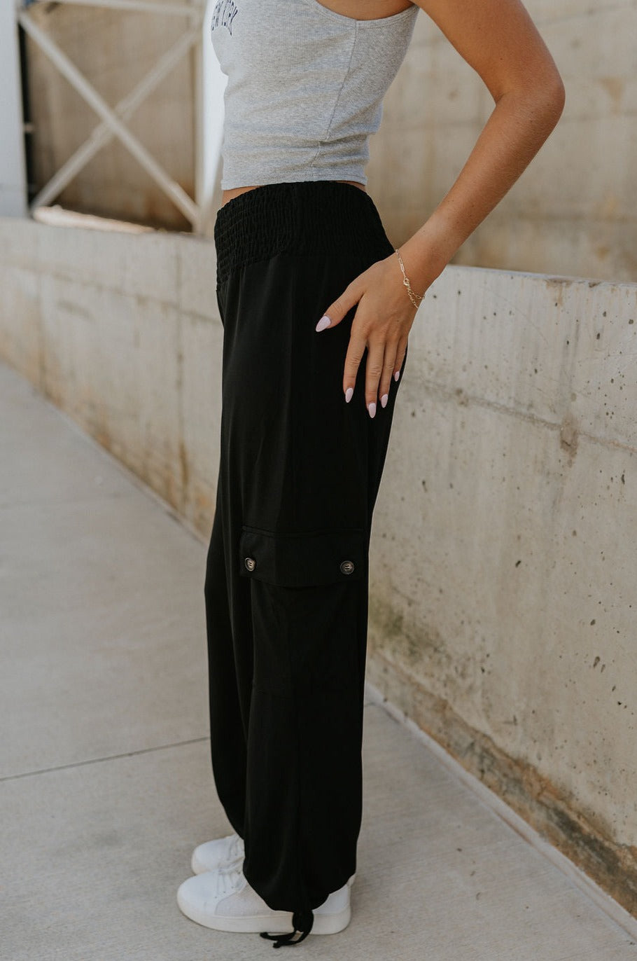 side view of female model wearing the Serena Black Cargo Drawstring Pants which features Black Lightweight Fabric, Jogger Pant Legs with Drawstring Ties, Cargo Pockets with Tortoise Buttons and Smocked Elastic Waistband
