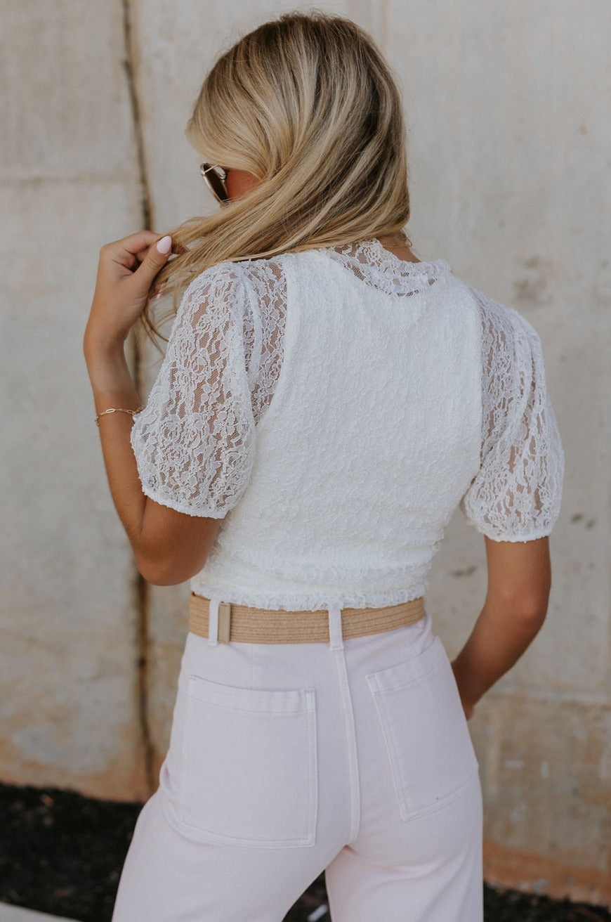 back view of female model wearing the Nylah Ivory Lace Short Sleeve Top which features White Lace Fabric, Lettuce Hem Details, Slight High Neckline and Short Puff Sleeves