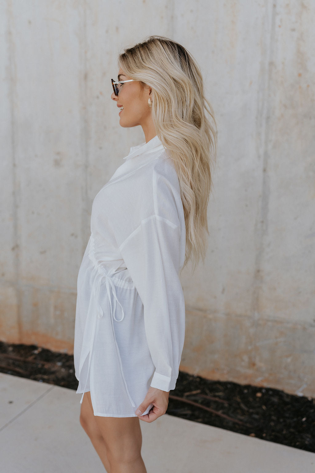 side view of female model wearing the Laura Off White Drawstring Cinched Long Sleeve Top which features Off White Lightweight Fabric, Slit Hem Details, Front Button-Up, Adjustable Cinch Waistband, Collared Neckline and Long Sleeves with Buttoned Cuffs