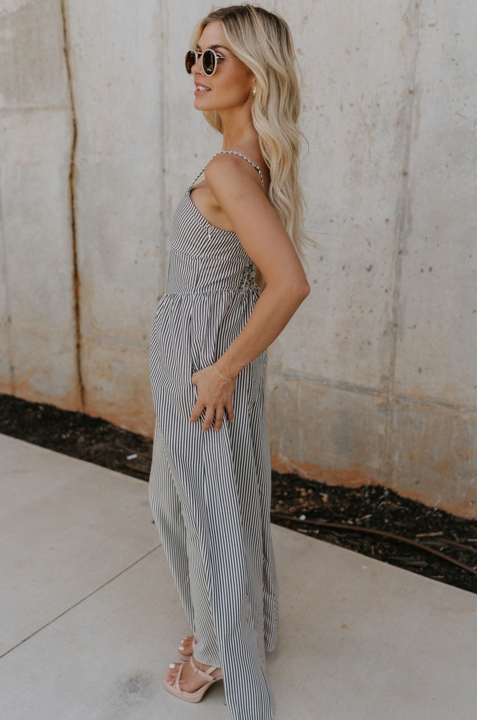side view of female model wearing the Palmer Black & Cream Stripe Sleeveless Jumpsuit which features Black and Cream Linen Stripe Pattern, Wide Pant Legs, Side Pockets, Sweetheart Neckline, Side Zipper, Adjustable Straps and Smocked Back