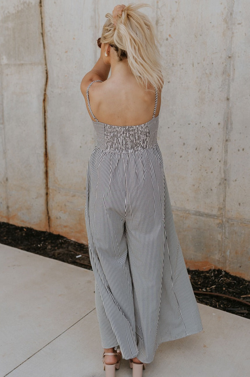 back view of female model wearing the Palmer Black & Cream Stripe Sleeveless Jumpsuit which features Black and Cream Linen Stripe Pattern, Wide Pant Legs, Side Pockets, Sweetheart Neckline, Side Zipper, Adjustable Straps and Smocked Back