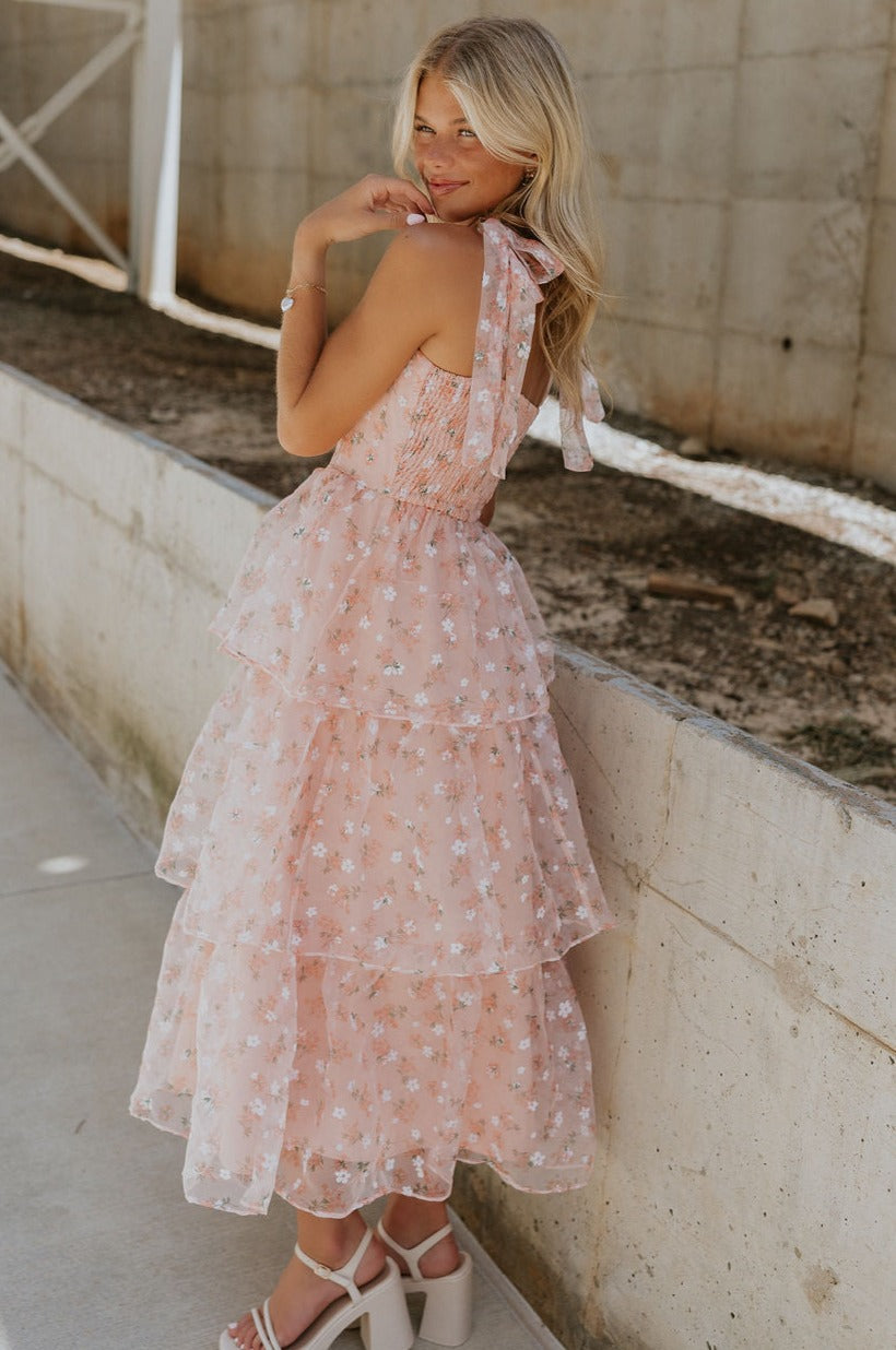side view of female model wearing the Evie Peach Blush Floral Tiered Midi Dress which features Light Peach Sheer Fabric, Floral Print, Tiered Design, Midi Length, Light Peach Lining, Square Neckline, Tie Straps and Smocked Back
