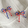 Image shows Quinn Red, White, & Blue Bow Earrings against a white background. Earrings have red white and blue beads in the shape of a bow, and the center is gold with rhinestones.