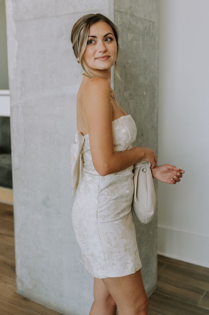 side view of female model wearing the Blakely Ivory & Gold Floral Bow Mini Dress which features Cream & Gold Jacquard Fabric, Floral Print, Mini Length, Cream Lining, Strapless, Back Bow Design nd Monochrome Side Zipper with Hook Closure