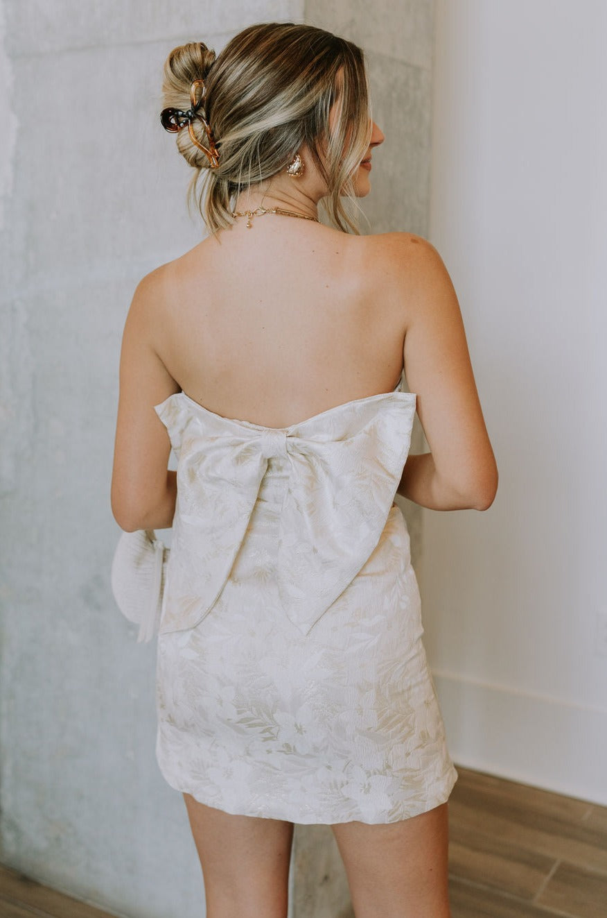 back view of female model wearing the Blakely Ivory & Gold Floral Bow Mini Dress which features Cream & Gold Jacquard Fabric, Floral Print, Mini Length, Cream Lining, Strapless, Back Bow Design nd Monochrome Side Zipper with Hook Closure