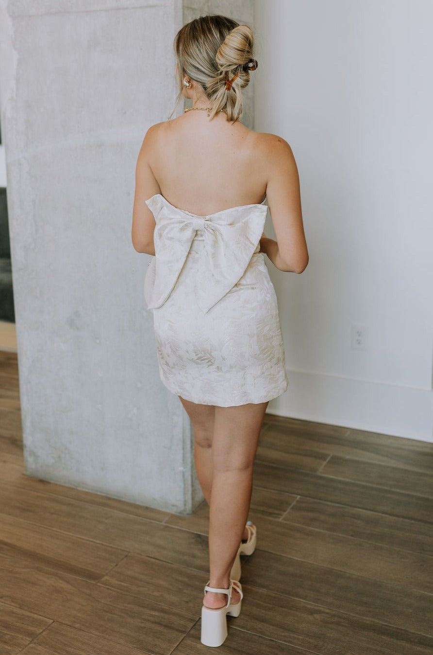 Full body back view of female model wearing the Blakely Ivory & Gold Floral Bow Mini Dress which features Cream & Gold Jacquard Fabric, Floral Print, Mini Length, Cream Lining, Strapless, Back Bow Design nd Monochrome Side Zipper with Hook Closure