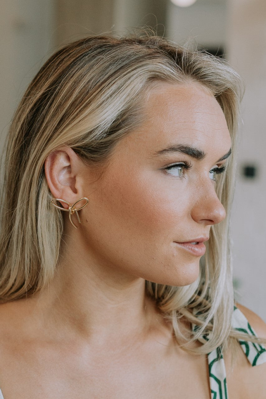 Side view of female model wearing the Charlie Gold Bow Shaped Stud Earring which features gold bow shaped studs