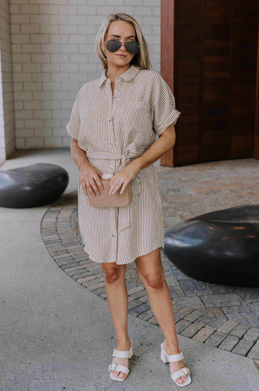 Full body front view of female model wearing the Valentina Taupe & Cream Striped Romper that has cream and taupe vertical stripes, a button up front with a collar, a tie belt, and short sleeves.