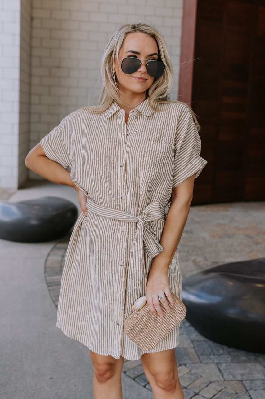 Upper body front view of female model wearing the Valentina Taupe & Cream Striped Romper that has cream and taupe vertical stripes, a button up front with a collar, a tie belt, and short sleeves.