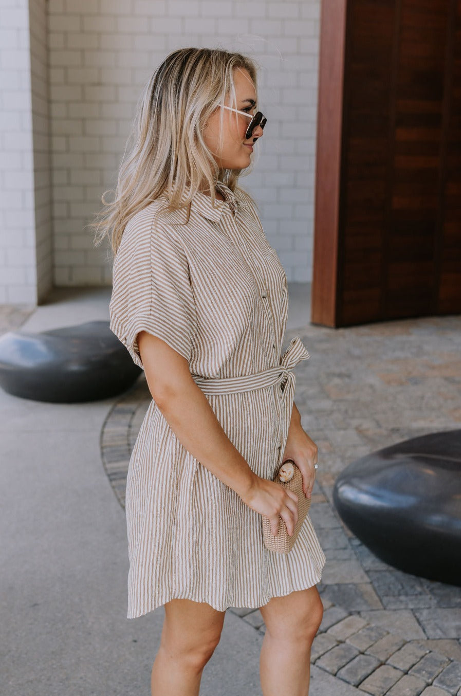 Side view of female model wearing the Valentina Taupe & Cream Striped Romper that has cream and taupe vertical stripes, a button up front with a collar, a tie belt, and short sleeves.