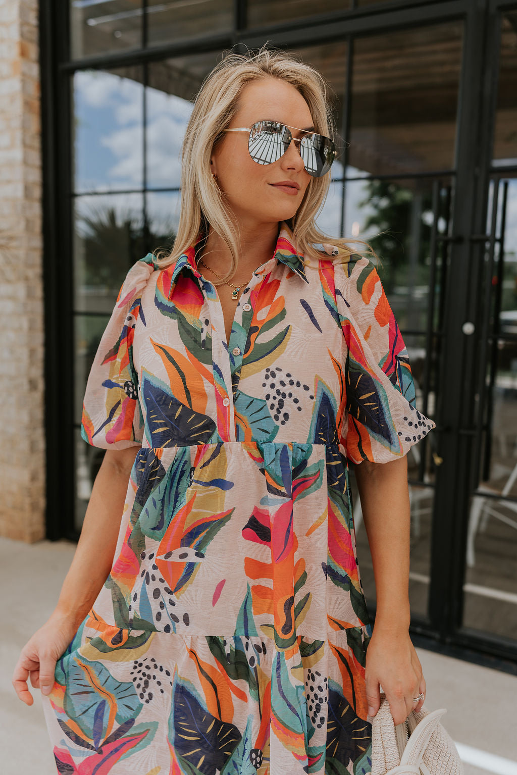 front view of female model wearing the Alora Multi Palm Floral Midi Dress which features Multi Color Floral Palm Print, Tan Lining, Tiered Body, Midi Length, Quarter Button-Up, Collared Neckline and Short Puff Sleeves