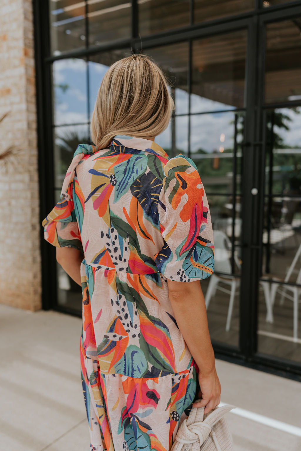 back view of female model wearing the Alora Multi Palm Floral Midi Dress which features Multi Color Floral Palm Print, Tan Lining, Tiered Body, Midi Length, Quarter Button-Up, Collared Neckline and Short Puff Sleeves