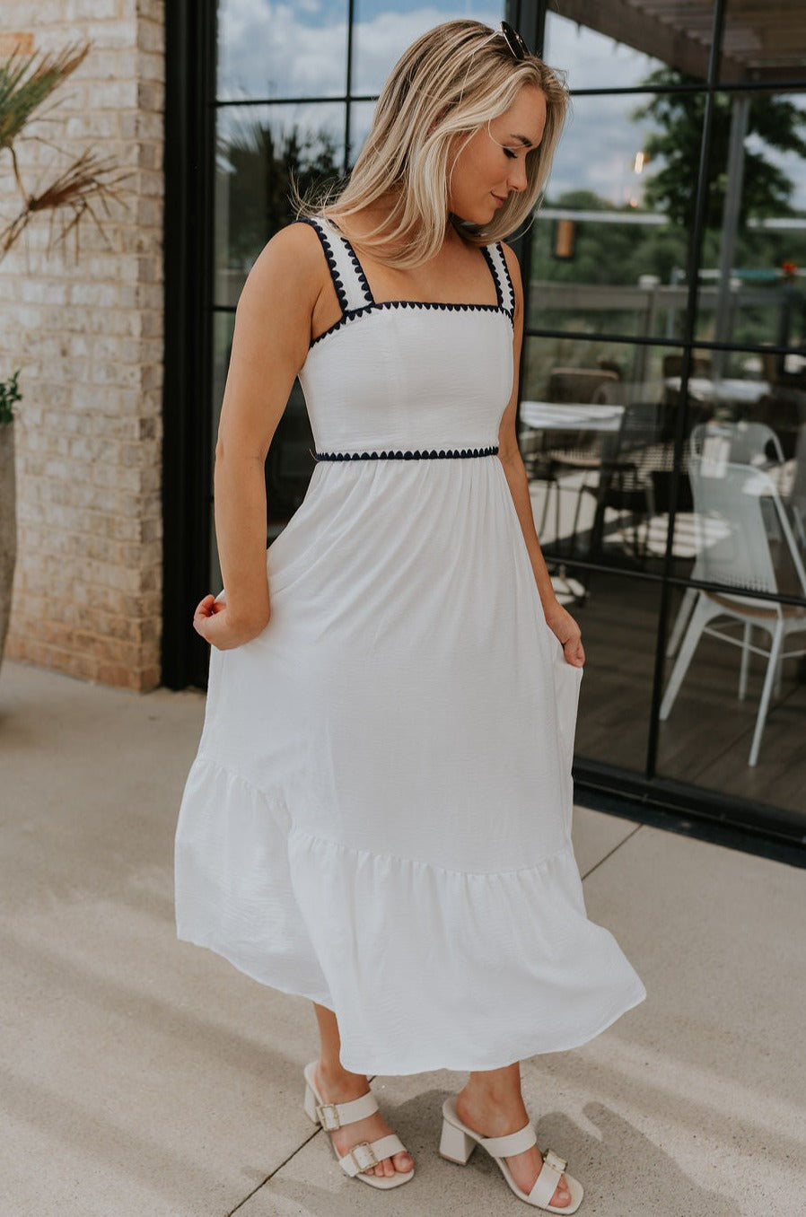 Full body side view of female model wearing the Saylor White & Navy Scallop Midi Dress which features White Lightweight Fabric, Navy Scallop Details, Peplum Hem, Midi Length, White Lining, Square Neckline and Monochrome Back Zipper with Hook Closure