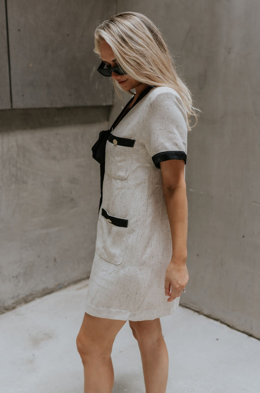 side view of female model wearing the Stevie Oatmeal & Black Short Sleeve Mini Dress which features Oatmeal Fabric, Black Trim Details, Mini Length, Anchor Button Up Detail, Four Buttoned Front Pockets, Black Tie Detail and Short Sleeves