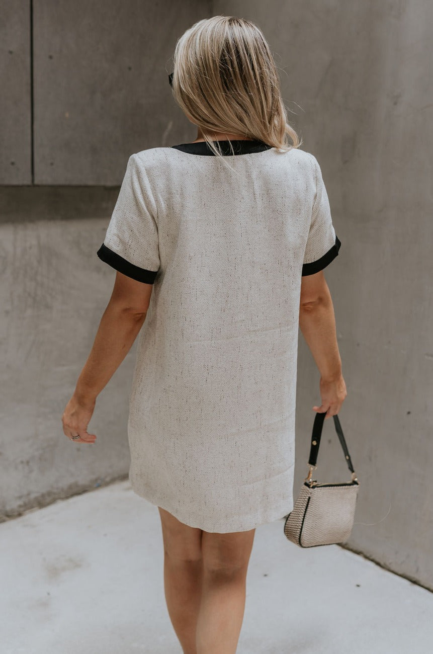 back view of female model wearing the Stevie Oatmeal & Black Short Sleeve Mini Dress which features Oatmeal Fabric, Black Trim Details, Mini Length, Anchor Button Up Detail, Four Buttoned Front Pockets, Black Tie Detail and Short Sleeves