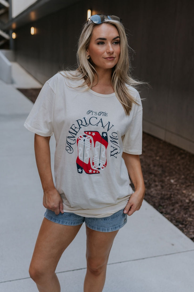 Full body view of female model wearing the It's The American Way Graphic Tee which features ream cotton fabric, round neckline, short sleeves and red, white and blue cowgirl boots graphic with "It's The American Way" writing.