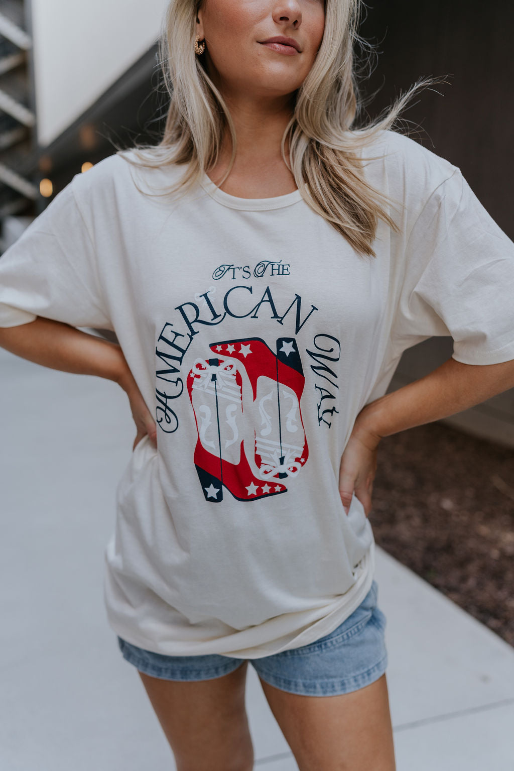 close up view of female model wearing the It's The American Way Graphic Tee which features ream cotton fabric, round neckline, short sleeves and red, white and blue cowgirl boots graphic with "It's The American Way" writing.