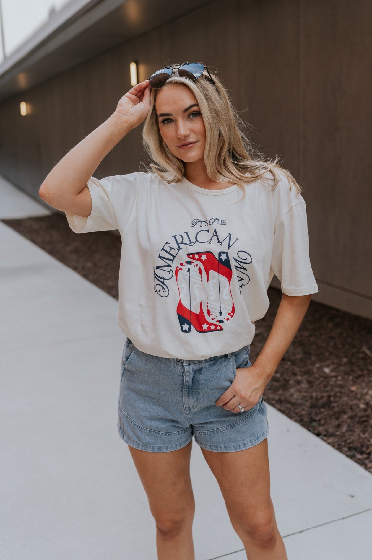 Full body view of female model wearing the It's The American Way Graphic Tee which features ream cotton fabric, round neckline, short sleeves and red, white and blue cowgirl boots graphic with "It's The American Way" writing.