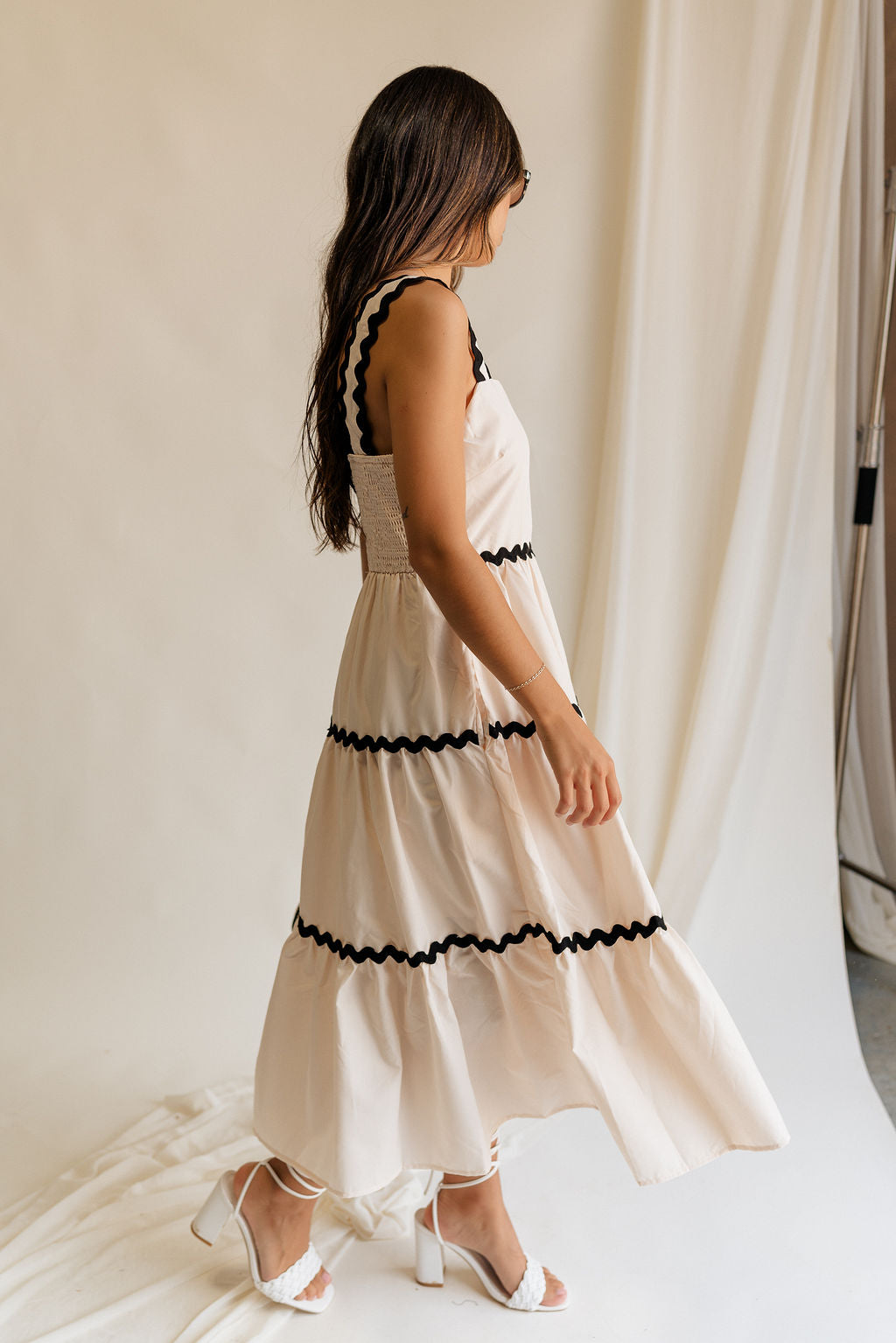 Full body side view of female model wearing the Clara Ecru & Black Wavy Trim Midi Dress which features Cream Lightweight Fabric, Cream Lining, Midi Length, Pockets on each side, Black Wavy Trim Details, Square Neckline, Straps and Smocked Back