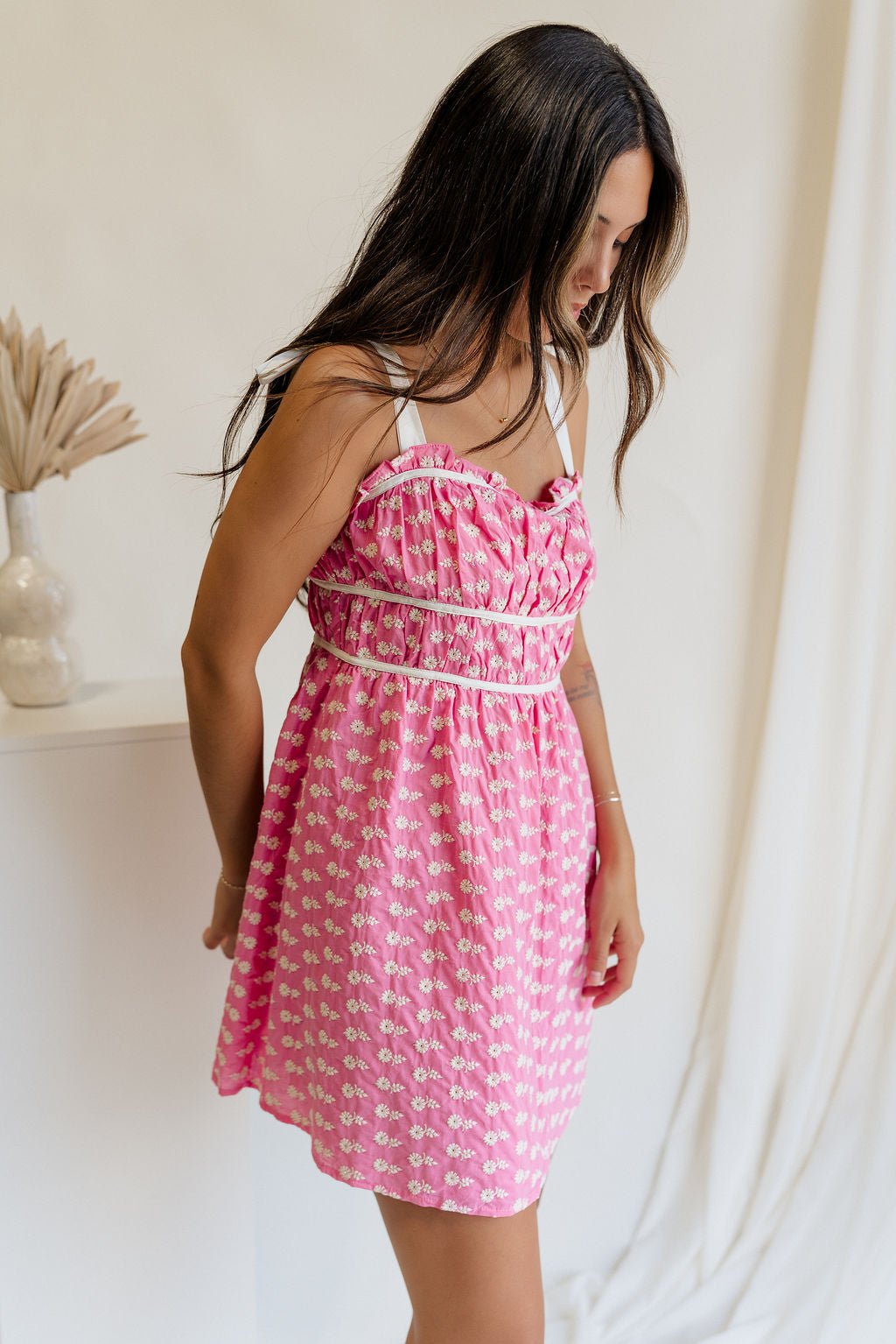 side view of female model wearing the Caroline Pink & White Floral Mini Dress which features Pink Lightweight Fabric, White Flower Pattern, Pink Lining, Ruffle Sweetheart Neckline, White Tie Straps and Monochrome Back Zipper with Hook Closure