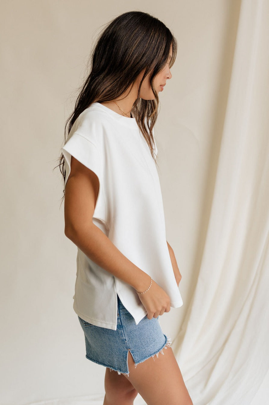 Side view of female model wearing the Blaire Off White Ribbed Short Sleeve Top which features Off White Ribbed Fabric, Round Neckline, Short Sleeves and Monochrome Button-Up Back Closure