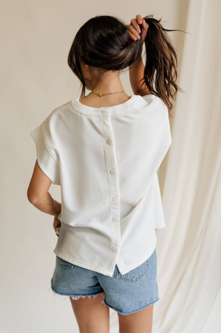 back view of female model wearing the Blaire Off White Ribbed Short Sleeve Top which features Off White Ribbed Fabric, Round Neckline, Short Sleeves and Monochrome Button-Up Back Closure