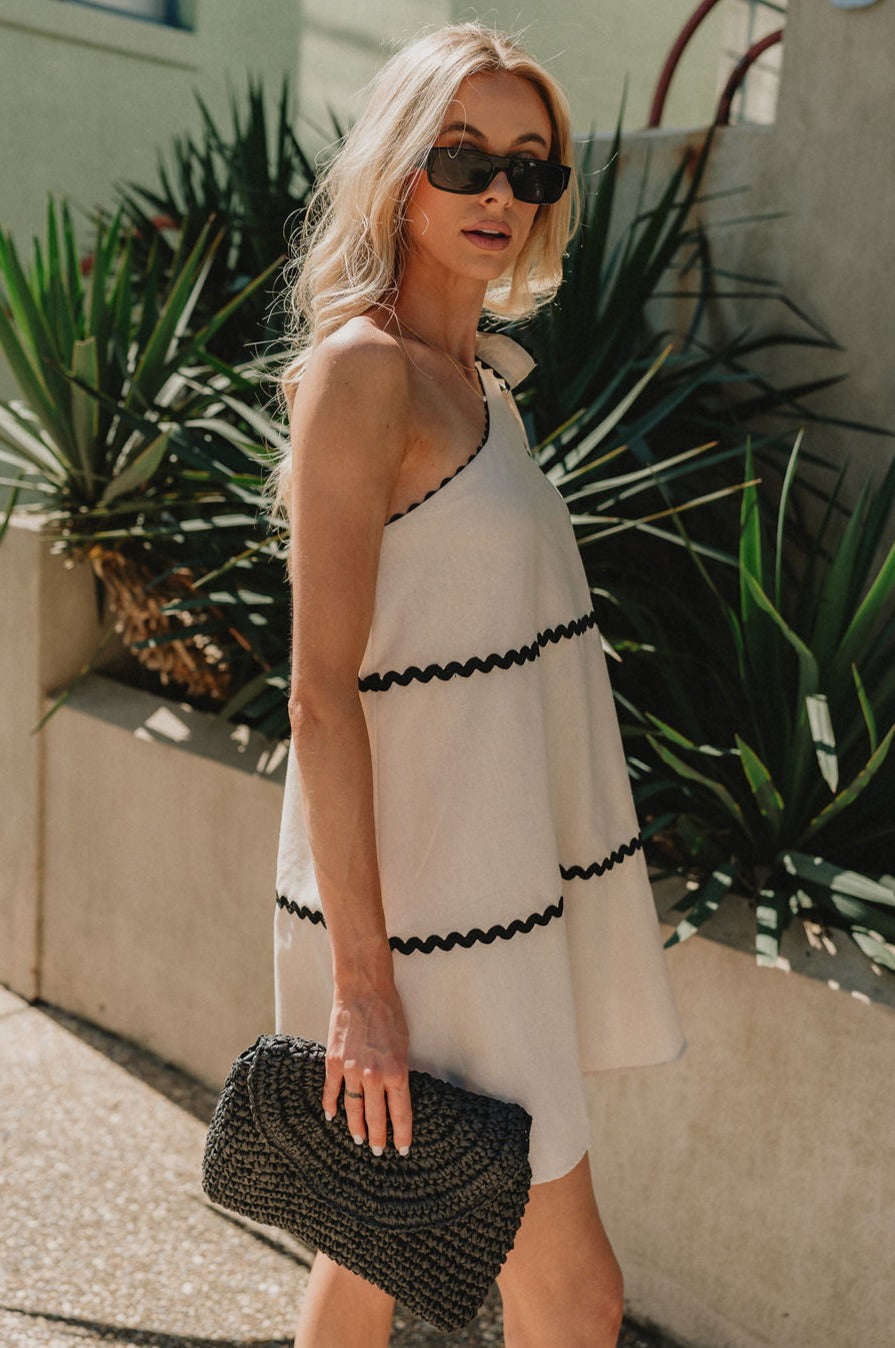 side view of female model wearing the Aria Oatmeal & Black Wavy One-Shoulder Mini Dress which features Cream Linen Lightweight Fabric, Cream Lining, Mini Length, Black Wavy Trim Details and One Shoulder Strap with Bow Tie