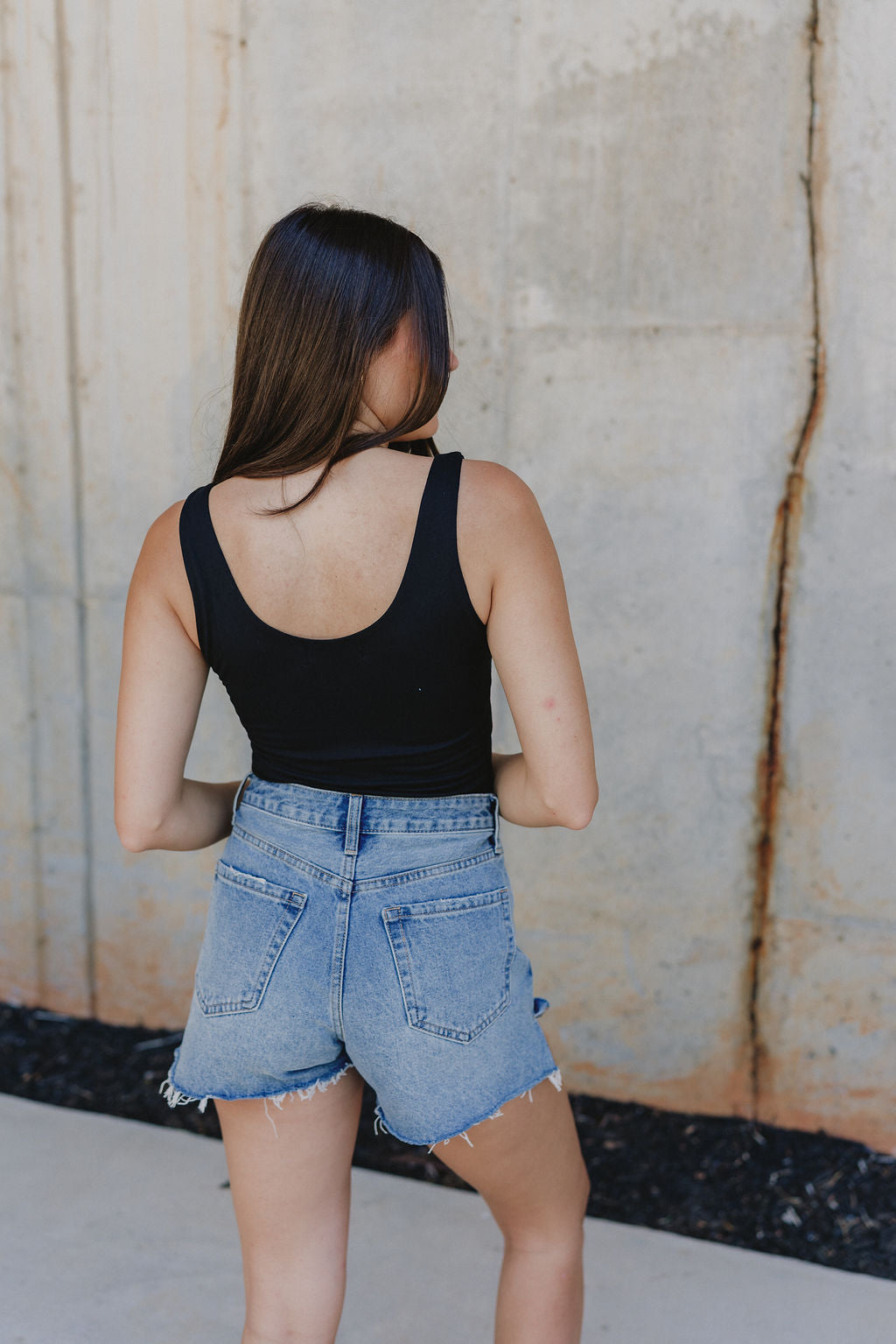 Back view of female model wearing the Mira Scooped Back Sleeveless Bodysuit which features a scoop neckline, Thick Straps, Scoop Back and Thong Bottom with Button Snap Closure.