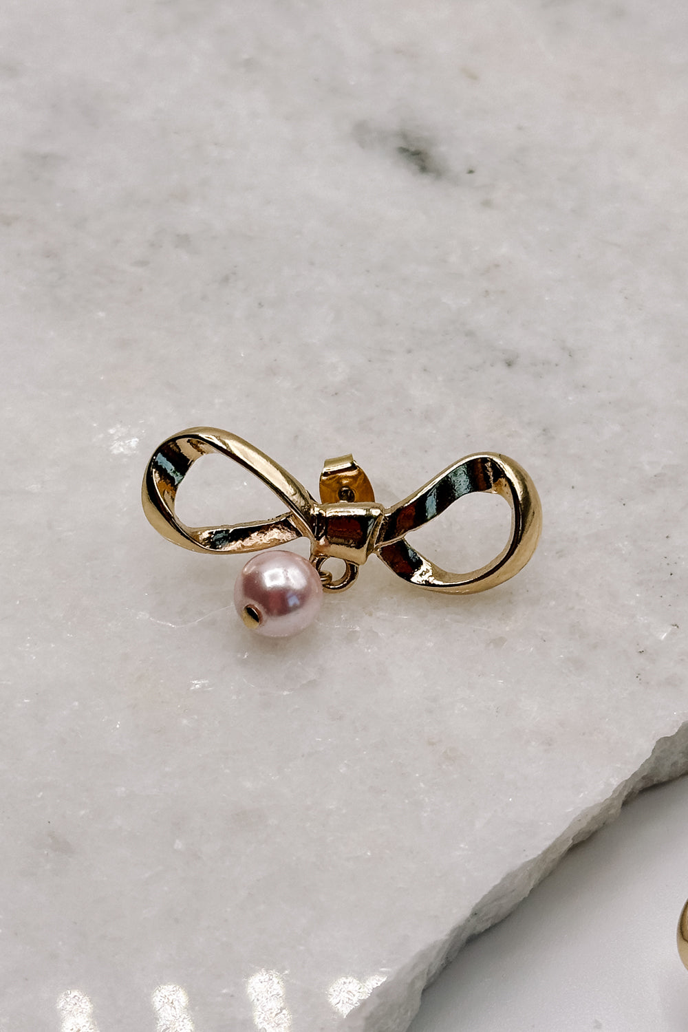 close up view of the Iris Pink & Gold Bow Stud Earring which features gold bow shaped studs with light pink pearl beads