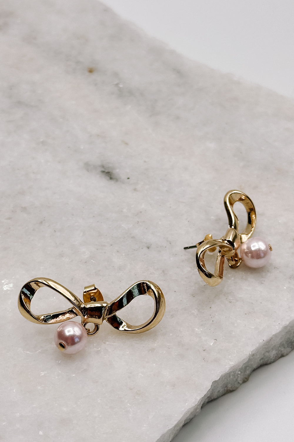 close up view of the Iris Pink & Gold Bow Stud Earring which features gold bow shaped studs with light pink pearl beads