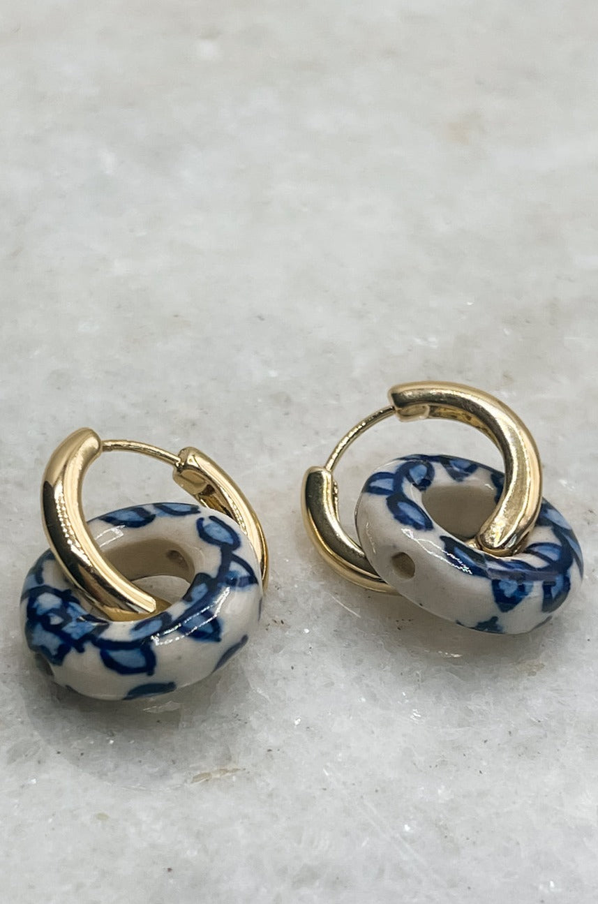 Close up of Lola White and Blue Pattern Link Earrings that are gold earrings with circle huggies that are a white and blue linked attachment