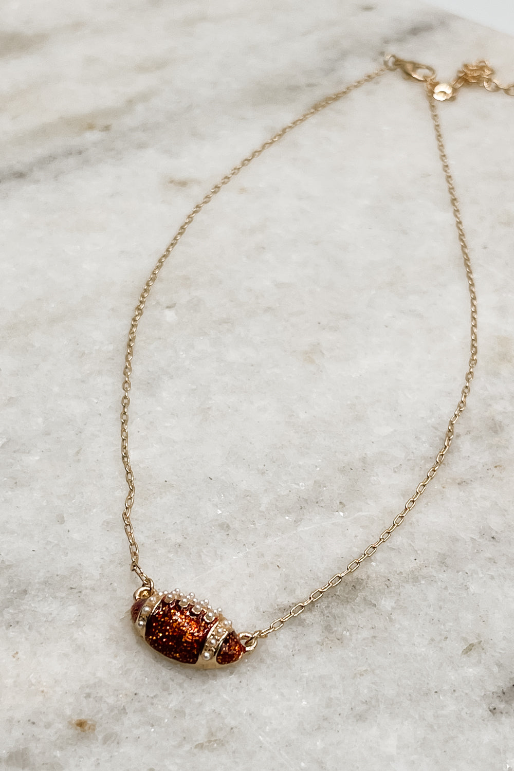 Image shows the Abby Gold Football Pendant Necklace against a marble background. Necklace features thin gold chain and a sparkly brown football pendant with pearl and gold detailing.