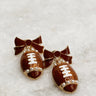 Photo shows a pair of earrings. Earrings are bown and gold bow studs with a bow. From the bow dangles a glittery brown football charm with pearl accents.
