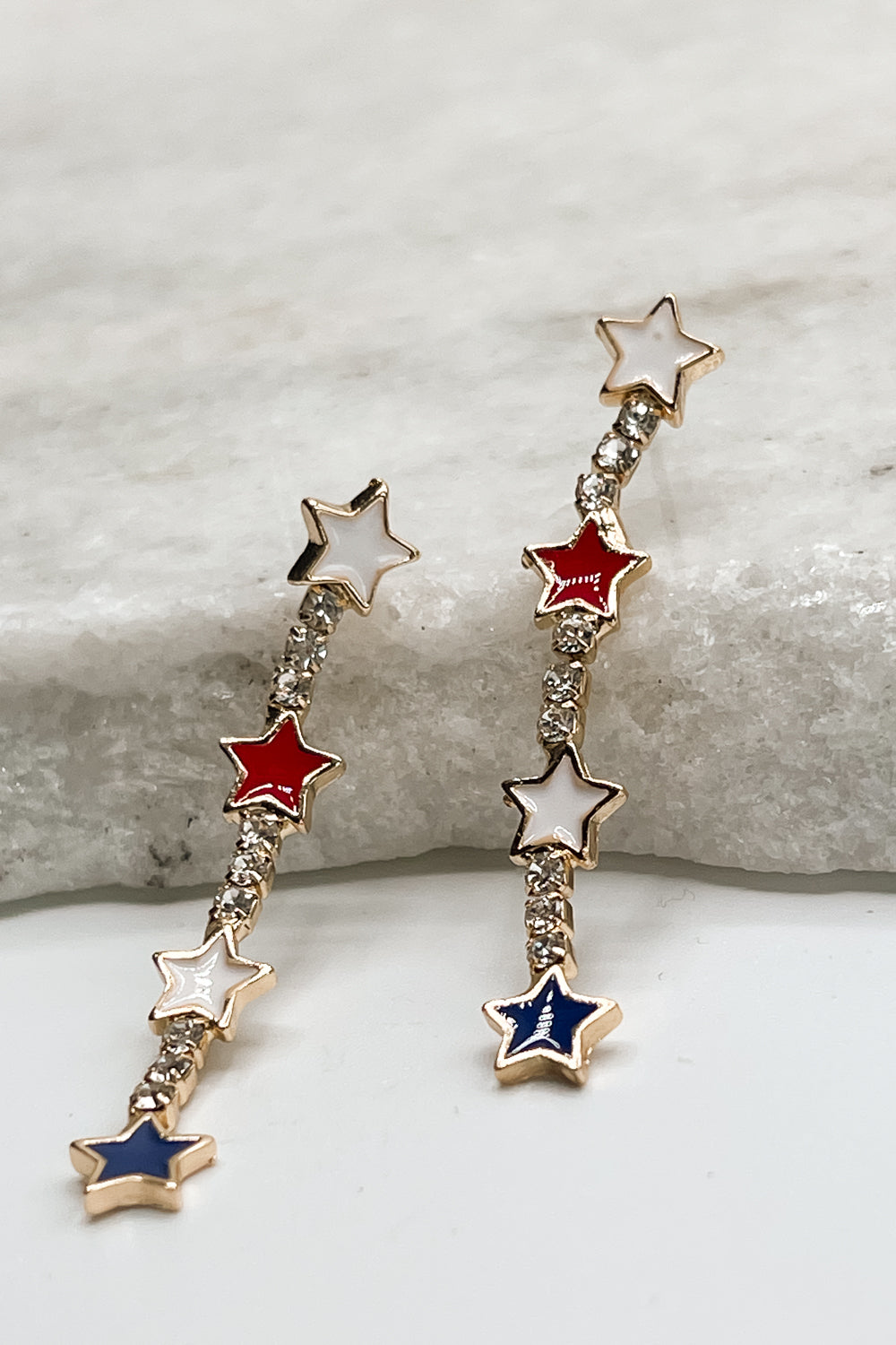 Image shows the Liberty Red, White, & Blue Star Earrings against a marble background. These earrings have 2 white stars, a red star, and a blue star in a vertical dangling line with rhinestones in between.