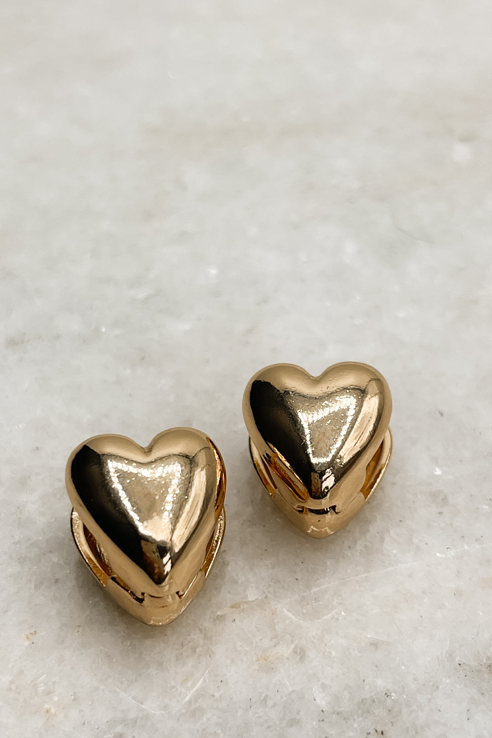 The Raine Earrings are shown against a neutral background. They are gold heart-shaped studs.