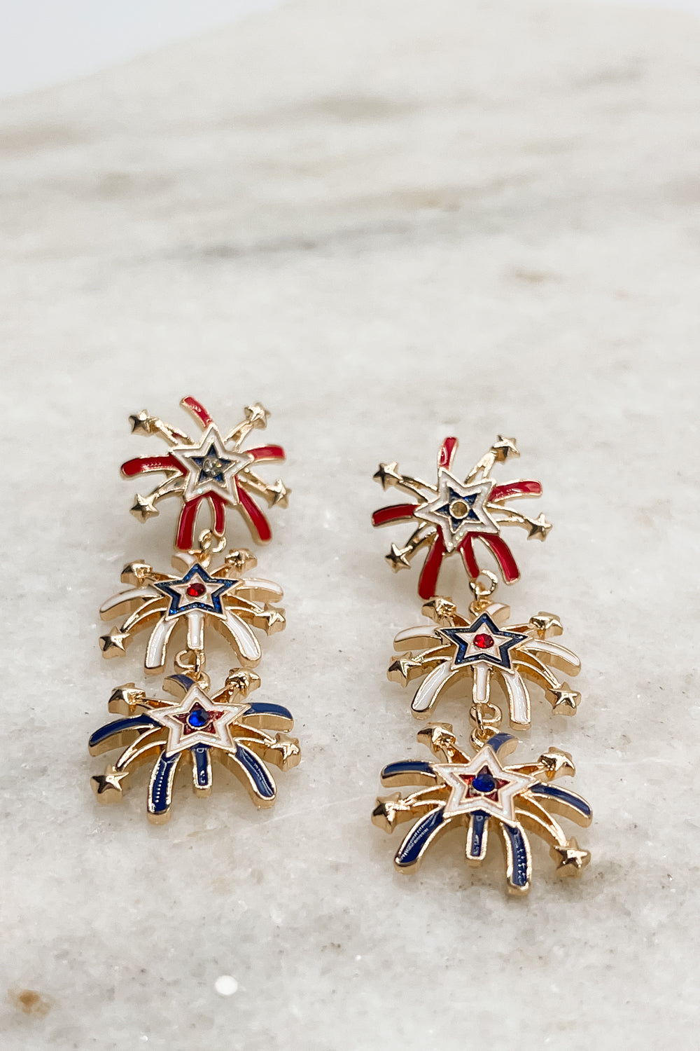 Earrings are shown straight-on lying on a neutral background. They are dangle earrings with three red, white, & blue firework-shaped charms.