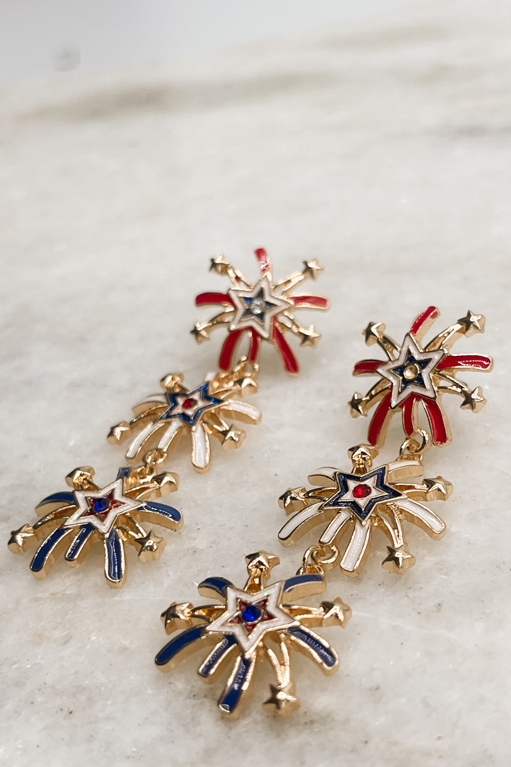 Earrings shown laid on a neutral background. They are dangle earrings with three red, white, & blue firework-shaped charms. 