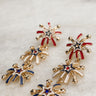 Earrings shown laid on a neutral background. They are dangle earrings with three red, white, & blue firework-shaped charms. 